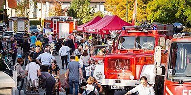 Menschenmenge beim Tag der offenen Tür der Abteilung Harthof mit roten Feuerwehrfahrzeugen und Pavillons in einer Straße mit herbstlichen Bäumen